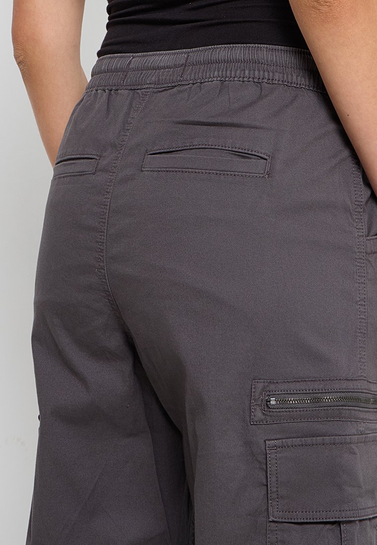 Szare spodnie cargo z elastycznym pasem. Posiadają dwie tylne kieszenie oraz boczną kieszeń na zamek. Materiał wygląda gładko, z matowym wykończeniem.