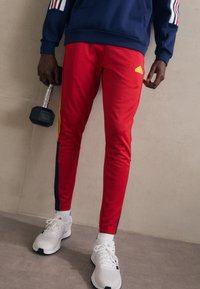 Pantaloni sportivi rossi con accenti gialli e strisce laterali blu, abbinati a una felpa navy e scarpe da ginnastica bianche, mentre tiene un manubrio.