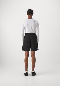 Vit långärmad skjorta, svarta shorts med elastisk midja, bakre logotypdetalj, bärs med svarta träningsskor mot en neutral bakgrund.