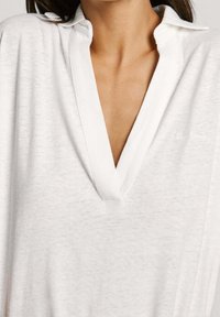 Vit tröja med djup V-ringning, tillverkad av lätt, texturerad tyg. Har en mjuk krage och subtil logotypdetalj längst ner till höger.