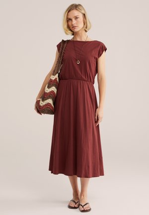 Nő bordó, ujjatlan midi ruhában, kerek nyakkivágással, fekete papucsban, medál nyaklánccal és nagy mintás válltáskával.