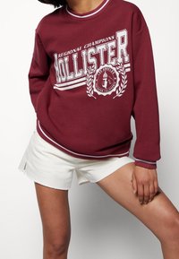 Sudadera burdeos con letras blancas, con la palabra "HOLLISTER" y un logo. Pantalones cortos blancos con dobladillo deshilachado. Ajuste casual y relajado.