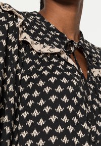 Blouse en tissu noir avec motif floral blanc ; possède un col à volants et une fermeture à nouer au niveau du cou. Texture lisse.