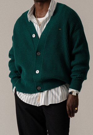 Mann trägt eine dunkelgrüne, gerippte Strickjacke über einem weißen Hemd mit schwarzen vertikalen Streifen, schwarze Hose, goldene Halskette, Ring und Armbanduhr.