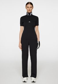 Sort kortærmet polo skjorte med højt krave og zebra mønster samt logo på brystet, parret med sorte træningsbukser og hvide sko.