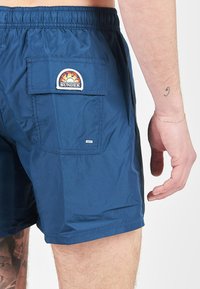 Bañador azul hecho de material ligero, con una cinturilla elástica y un bolsillo trasero con un parche de logo redondo.