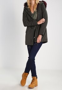 Olivgrüner Parka mit einer Kapuze aus Kunstfell, mit Reißverschluss und Druckknöpfen, kombiniert mit dunkelblauen Jeans und hellbraunen Stiefeletten.