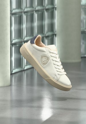 Basket blanche avec semelle beige et empiècement bleu marine au talon, en suspension dans un espace moderne et minimaliste aux murs en briques de verre.