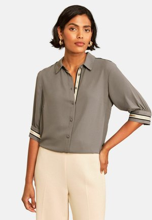 Woman wearing a gray button-up blouse with striped cuffs and cream-colored pants, standing with one hand on her hip against a plain background.