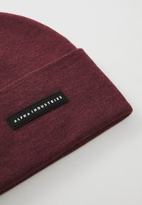 Burgundská pletená čepice s ohrnutým lemem, na kterém je černý obdélníkový štítek s nápisem "ALPHA INDUSTRIES" v bílém písmu.