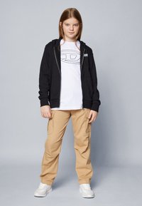 Felpa nera con zip, t-shirt bianca con grafica, pantaloni cargo beige e sneakers bianchi. Caratteristiche includono tasche e elementi di design casual.