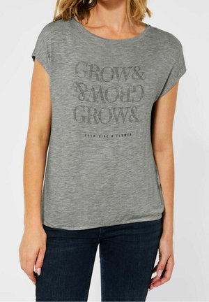 Kvinde iført en grå kortærmet skjorte med teksten "GROW & GROW & GROW" og "GROW LIKE A FLOWER" kombineret med mørke jeans.