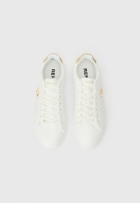 Sneakers brancos em couro com detalhes em dourado, apresentando uma superfície texturizada, design com atacadores e um logotipo na lateral. Atacadores e palmilha em dourado.