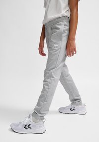 Graue Sweatpants mit einem elastischen Bund, Seitentaschen und einem weißen Logo auf der Hüfte. Kombiniert mit weißen Sportschuhen mit schwarzen Akzenten.