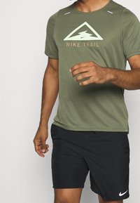 Grön kortärmad sporttröja med en strukturerad tyg, prydd med en vit och orange logotyp samt triangulärt mönster. Bärs med svarta shorts.