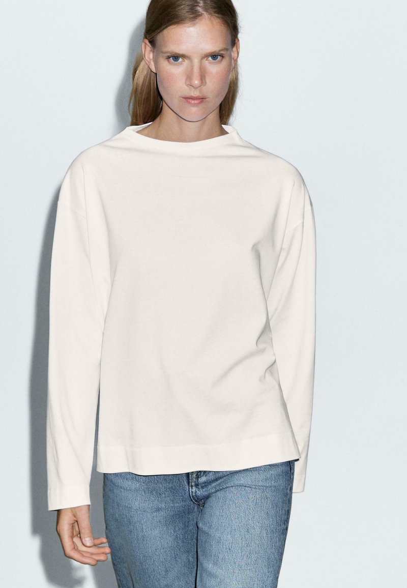 Kvinde med glat lyst brunt hår iført en ensfarvet hvid langærmet skjorte og lyseblå jeans mod en lysegrå baggrund.