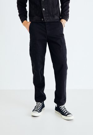 Czarna kurtka dżinsowa z guzikami, zestawiona z luźnymi czarnymi spodniami cargo z wieloma kieszeniami i mankietami. Całość uzupełniają czarne sneakersy typu high-top.