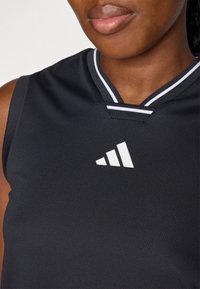Maillot de sport noir avec un col en V et des accents blancs. Il présente un logo Adidas blanc bien en vue sur le côté gauche de la poitrine. Tissu texturé.