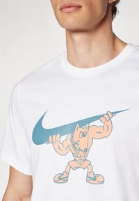Hvid bomulds t-shirt med et stort turkist Nike swoosh og en orange tegneseriefigur, der løfter vægte, med muskuløse træk.