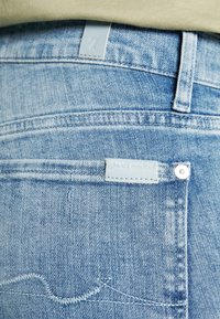 Jean denim bleu clair avec poche arrière, petite étiquette en cuir et passants de ceinture, porté avec un haut beige.