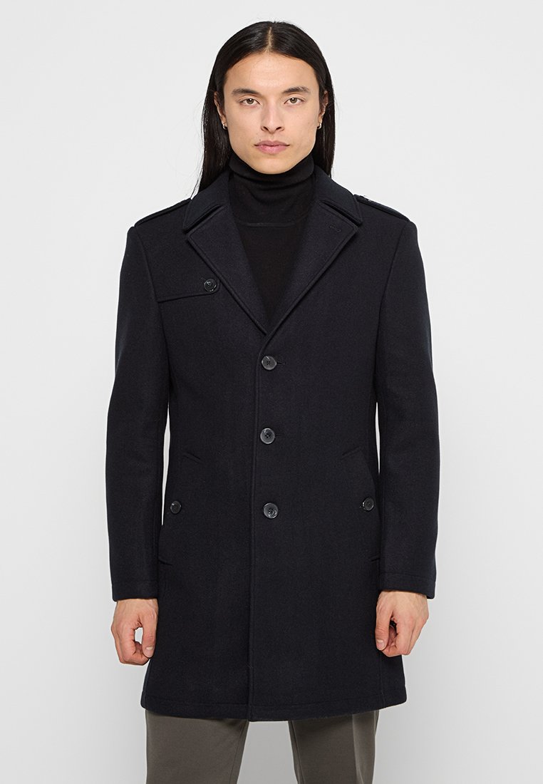 Cappotto in misto lana nera con design doppiopetto, grande colletto, tasca sul petto e due tasche fronte. I bottoni frontali sono tondi e neri.