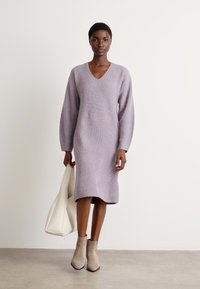 Trikotāžas, gara piedurkne, lilā kleita ar V veida izgriezumu, taisnu siluetu, ribotu tekstūru un sānu šķēlumiem, apvienota ar beigu potīšu zābakiem un baltu somu.