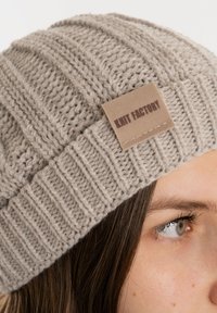 Gebreide muts in lichtgrijs met een ribbelstructuur. Heeft een tan leren label met "KNIT FACTORY" in reliëf, wat het ontwerp accentueert.