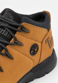 Boots Timberland în nuanțe de bej și negru, cu sireturi negre, guler căptușit și logo-ul cu copacul embosat pe lateral.