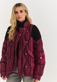 Burgundský prešívaný kabát s čiernymi textúrovanými rukávmi, obsahujúci vrecká, cvoky a zipsové zapínanie a detail v podobe motýlieho prívesku.