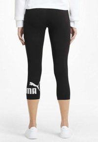 Osoba w czarnych, krótkich legginsach z białym logo Pumy na lewym łydce i białych tenisówkach, stojąca na jednolitym tle.