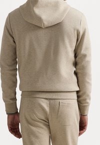 Ljusbeige hoodie i mjukt tyg med avslappnad passform, huva och ribbade muddar. Bakifrån syns en enkel design utan dekorationer.