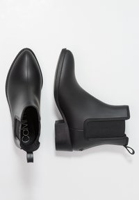 Bottines noires à bout pointu, en matière caoutchouteuse, avec des panneaux latéraux élastiques et un petit talon. Texture lisse avec peu de détails.