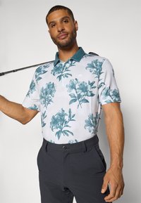 Kortsleeve polo skjorte i hvit med teal blomstermønster, ensfarget teal krage og logo detalj. Matchet med mørke bukser. Golfkølle holdt over skulderen.