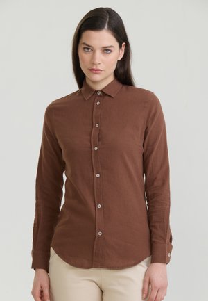 Femme aux longs cheveux foncés portant une chemise marron à manches longues boutonnée et un pantalon beige clair, debout devant un fond gris uni.