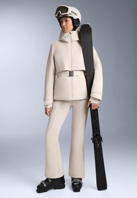 Skijaška odjeća uključuje skraćenu bež jaknu s remenom i odgovarajuće hlače, crne skijaške čizme i crnu skiju, uz bijelu kacigu i naočale za skijanje.