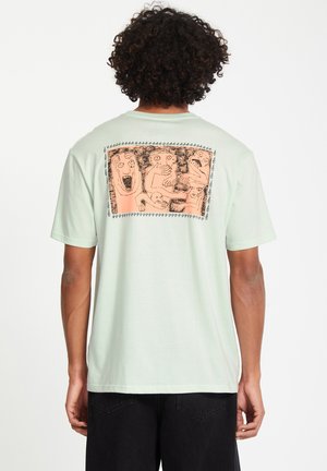 Lys grøn bomulds t-shirt med et stiliseret orange grafisk motiv på ryggen, der viser abstrakte figurer med sort kant. Korte ærmer.