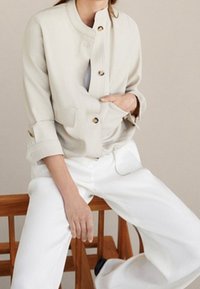 Beige cropped jakke med stående krave, guld knapper, to forlommer og oprullede ærmer, parret med brede hvide bukser.