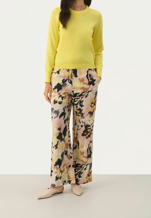 Femme portant un haut jaune vif à manches longues, un pantalon large à motif floral dans des tons roses, noirs et jaunes, et des chaussures plates couleur nude.