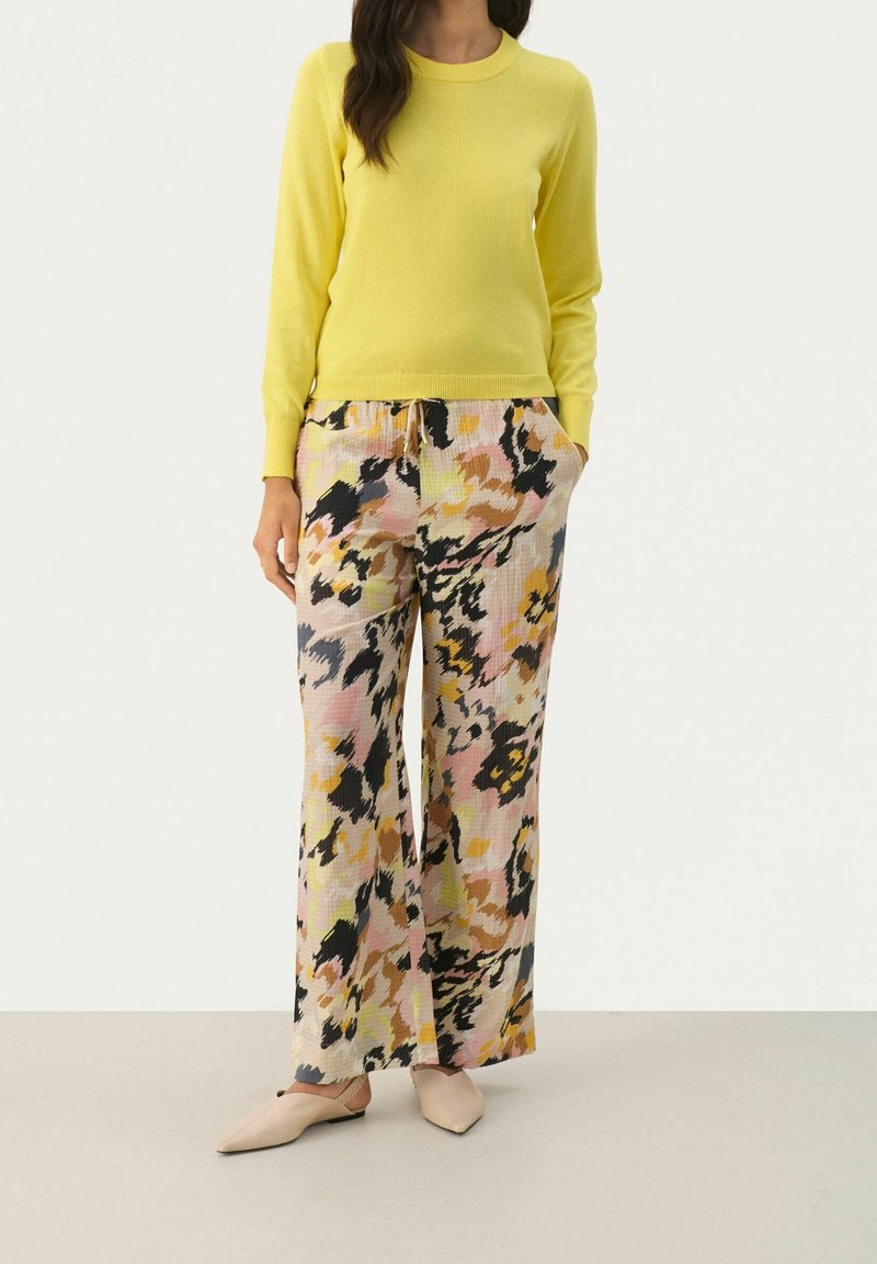 Femme portant un haut jaune vif à manches longues, un pantalon large à motif floral dans des tons roses, noirs et jaunes, et des chaussures plates couleur nude.