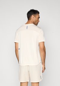 Light beige athletic shirt with short sleeves, smooth texture, and dotted accents in navy blue on the back and side, paired with matching shorts.