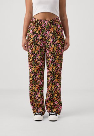 Person iført løstsiddende sorte bukser med blomsterprint i pink, orange og gul, hvid crop top og hvide sneakers med sorte snørebånd.