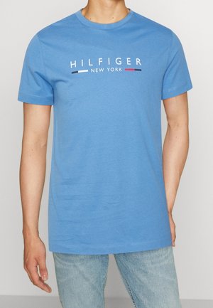 Man wearing a blue short-sleeve T-shirt with "Hilfiger New York" text and red, white, and blue stripes, paired with light blue jeans.