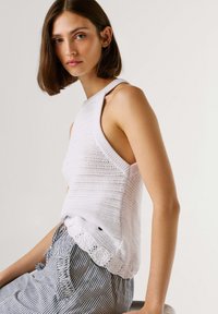 Jonge vrouw met kort bruin haar, gekleed in een witte, gebreide mouwloze top en gestreepte, hoog getailleerde broek, zittend tegen een effen achtergrond.