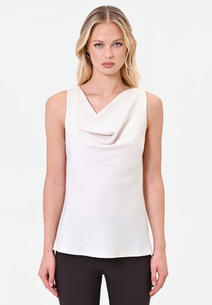 Blonde woman wearing sleeveless white top with draped neckline and black pants, standing against plain light background.