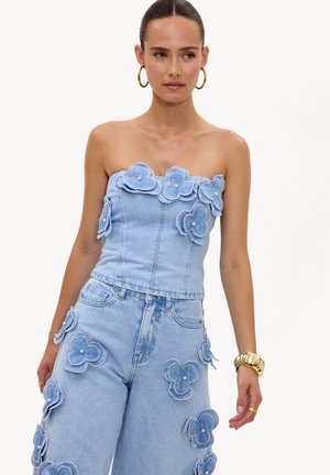 Kobieta ubrana w jasnoniebieski bezramiączkowy top i spodnie z denimu, ozdobione dużymi kwiatami z materiału z perłowymi środkami, złote kolczyki-koła i bransoletka.