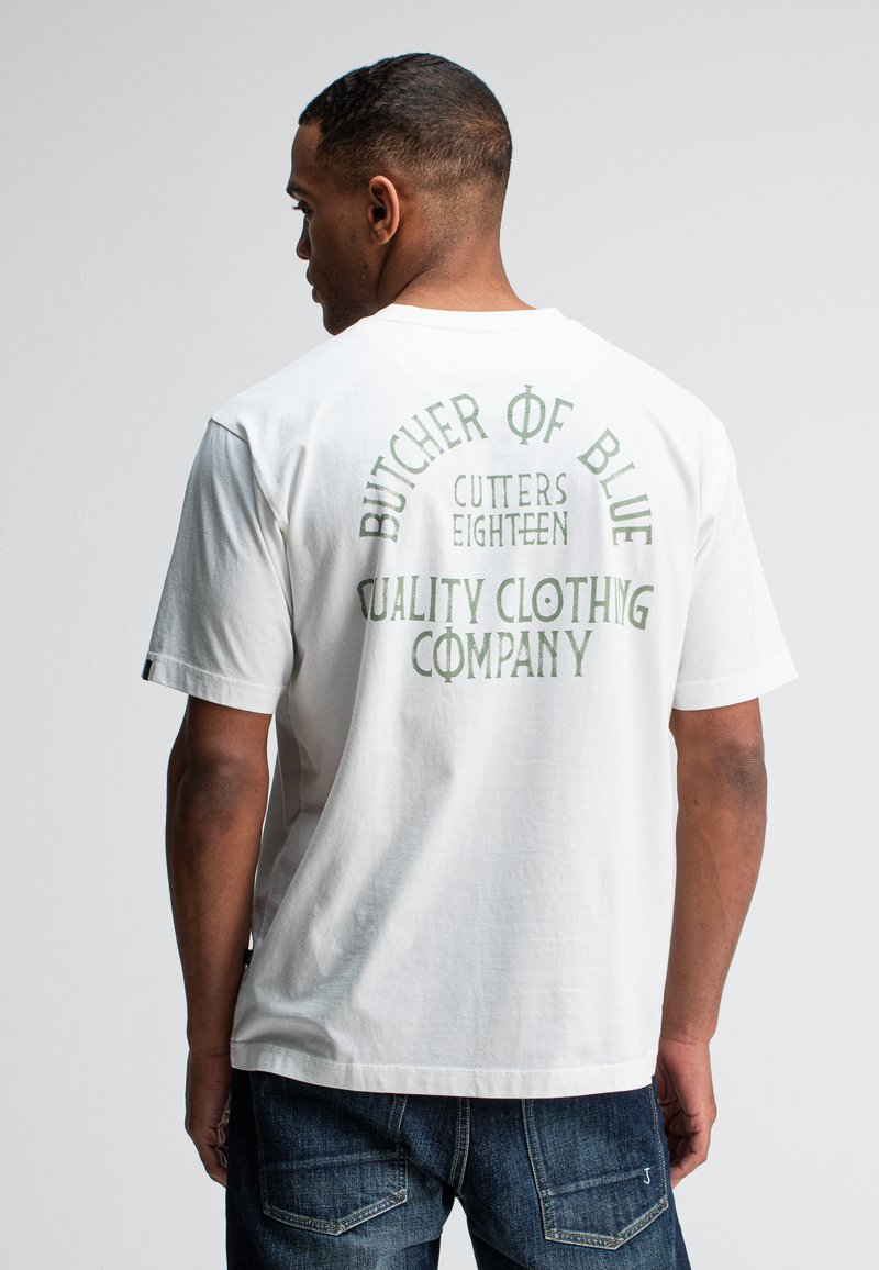 Mann in weißem T-Shirt mit grünem Text "Butcher of Blue Cutters Eighteen Quality Clothing Company" auf dem Rücken, kombiniert mit blauen Jeans.