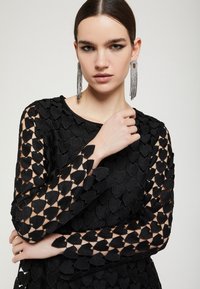 Mustassa mekossa on pitkät hihat ja sydämen muotoinen leikkaus. Mallilla on pitkät hopeiset frangee korvakorut. Materiaali on sileä.