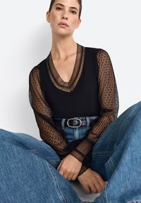 Zwarte top met doorschijnende, patroon lange mouwen, V-hals, gecombineerd met hoge taille blauwe denim jeans en een zwarte riem in de taille.