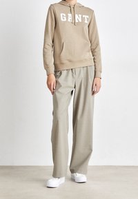 Béžová mikina s bílým nápisem "GANT", v kombinaci se světle šedými kalhotami s širokými nohavicemi a bílými teniskami, na neutrálním pozadí.