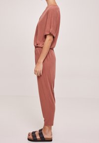 Jumpsuit rose avec des manches courtes, ceinture élastique et jambes fuselées, associé à des sandales à plateforme noires. Texture douce, coupe décontractée.